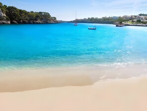 A beach with clear turquoise water, a sandy shore, and a boat in the distance. - Villas2meet Son Rapinya Porto Cristo (Manacor)