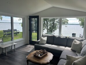 A modern living room with a grey sofa, a wooden coffee table, and a bench by a large window overlooking a lake.