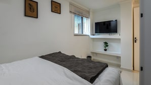 A bedroom with a bed, a television on a shelf, a window with blinds, and a small plant on a shelf. - ApartDeck Fantasy Island (Rio de Janeiro)