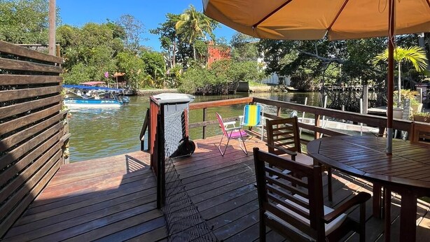 A wooden deck with a table and chairs, a covered area with an umbrella, and a view of a river with boats. - ApartDeck Fantasy Island (Rio de Janeiro)