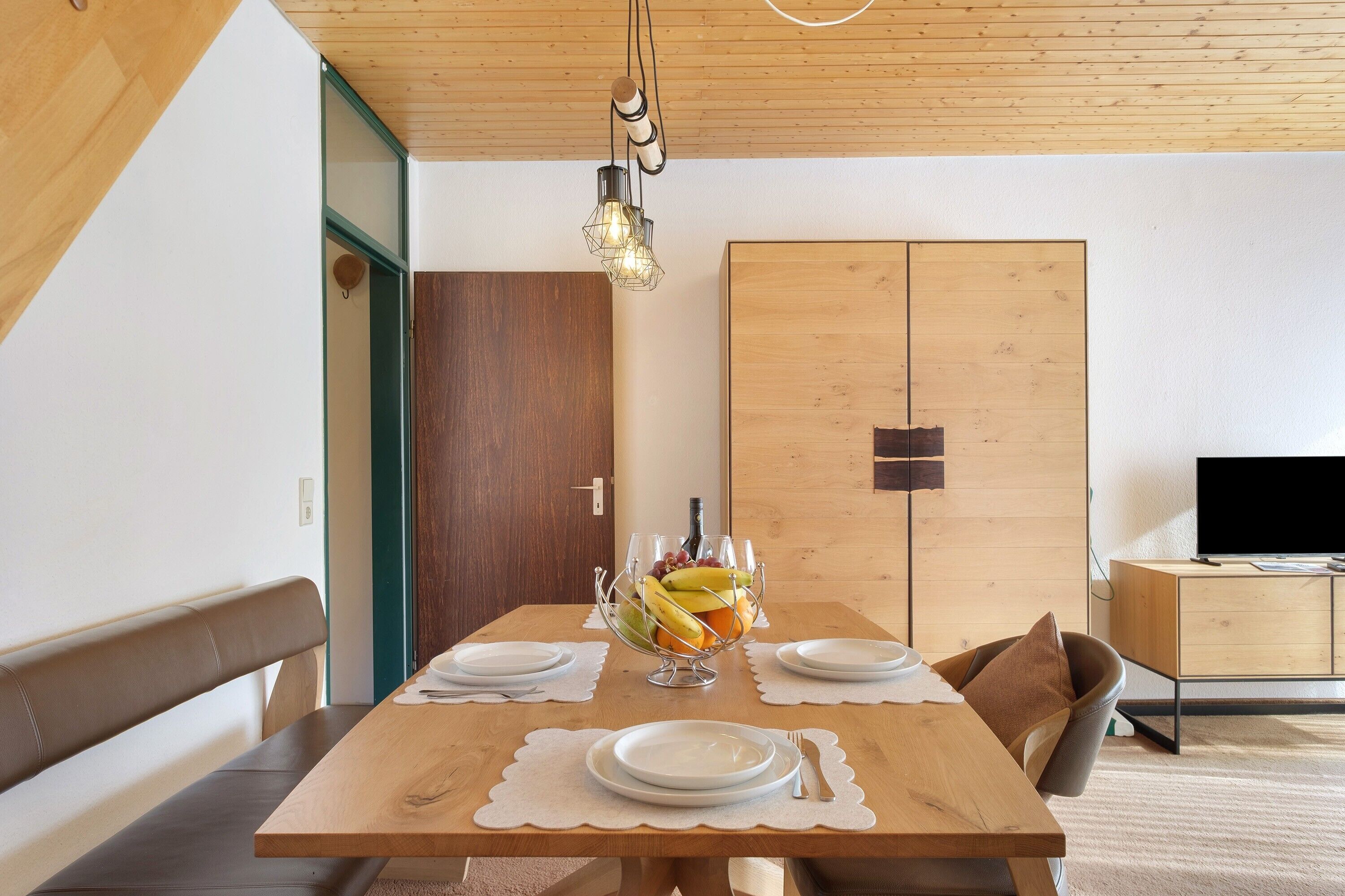 A dining table with a fruit bowl, plates, and cutlery. A wooden bench and a television are visible in the background.
