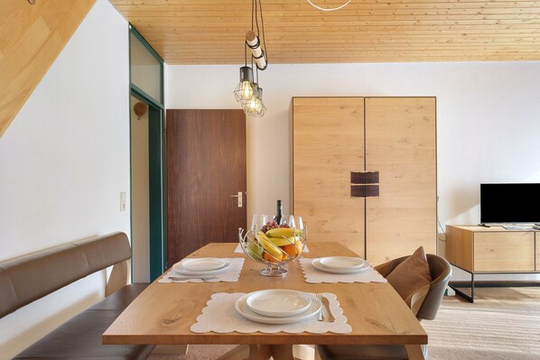 Une table à manger avec un panier de fruits, des assiettes et des couverts. On aperçoit un banc en bois et un téléviseur à l’arrière-plan.