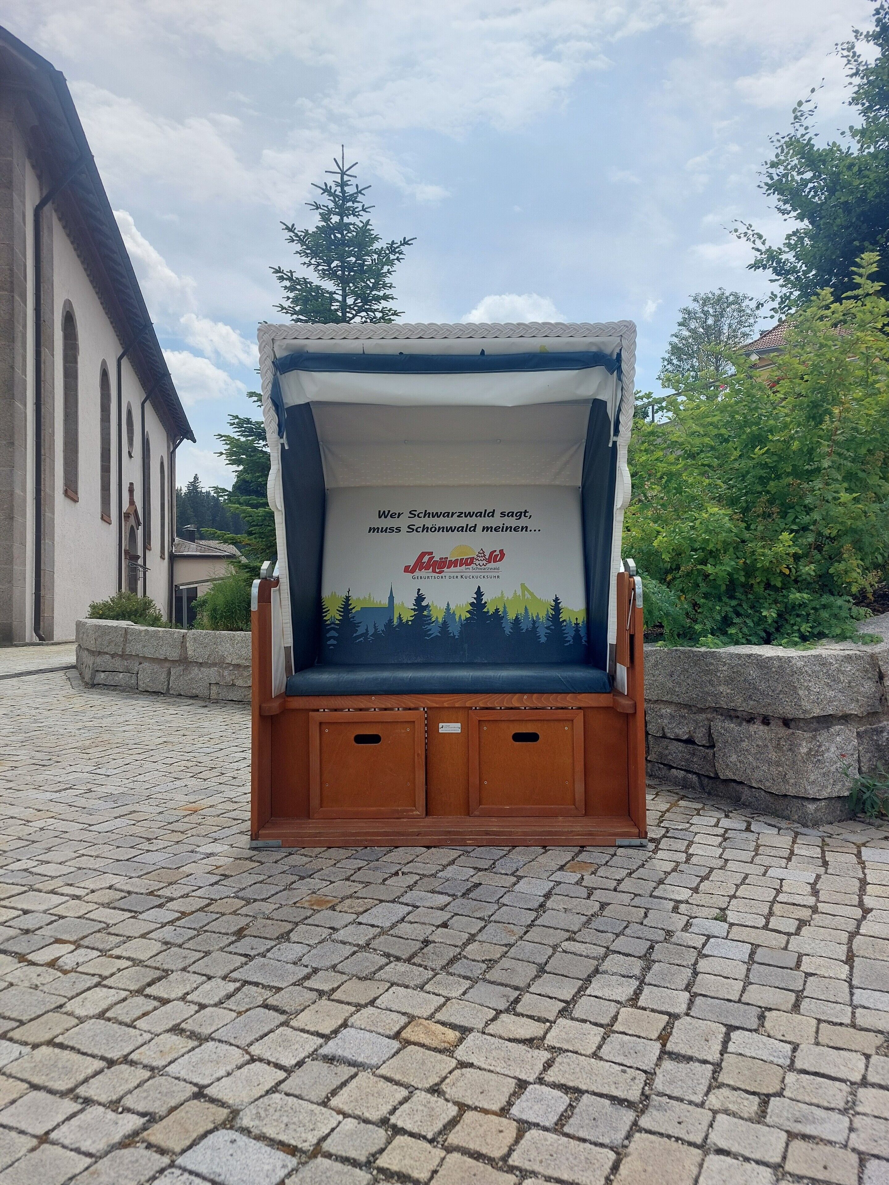 Eine Holzbank mit Rückenlehne und einer aufgedruckten Nachricht vor einem Gebäude.