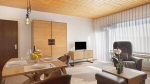 A modern living room with a wooden ceiling, a dining table set with plates and a fruit bowl, a leather armchair, and a television on a wooden cabinet.
