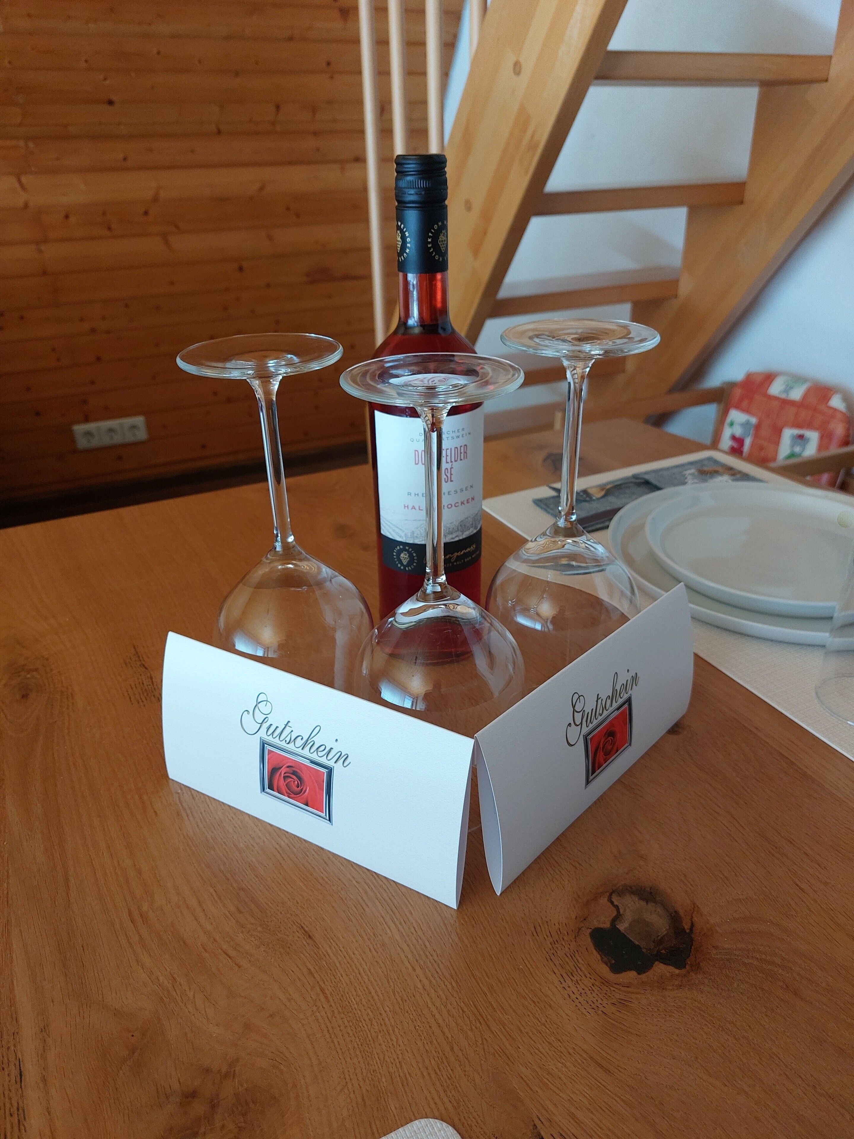 Une table en bois avec une bouteille de vin rouge, trois verres à vin et deux boîtes blanches étiquetées « Gutschein ».