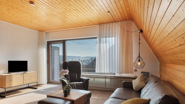 A modern living room with a wooden ceiling, a large window with curtains, a leather sofa, a coffee table, and a TV on a wooden cabinet.