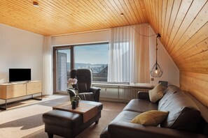 Un salon moderne avec un plafond en bois, une grande fenêtre avec des rideaux, un canapé en cuir, une table basse et un téléviseur posé sur un meuble en bois.