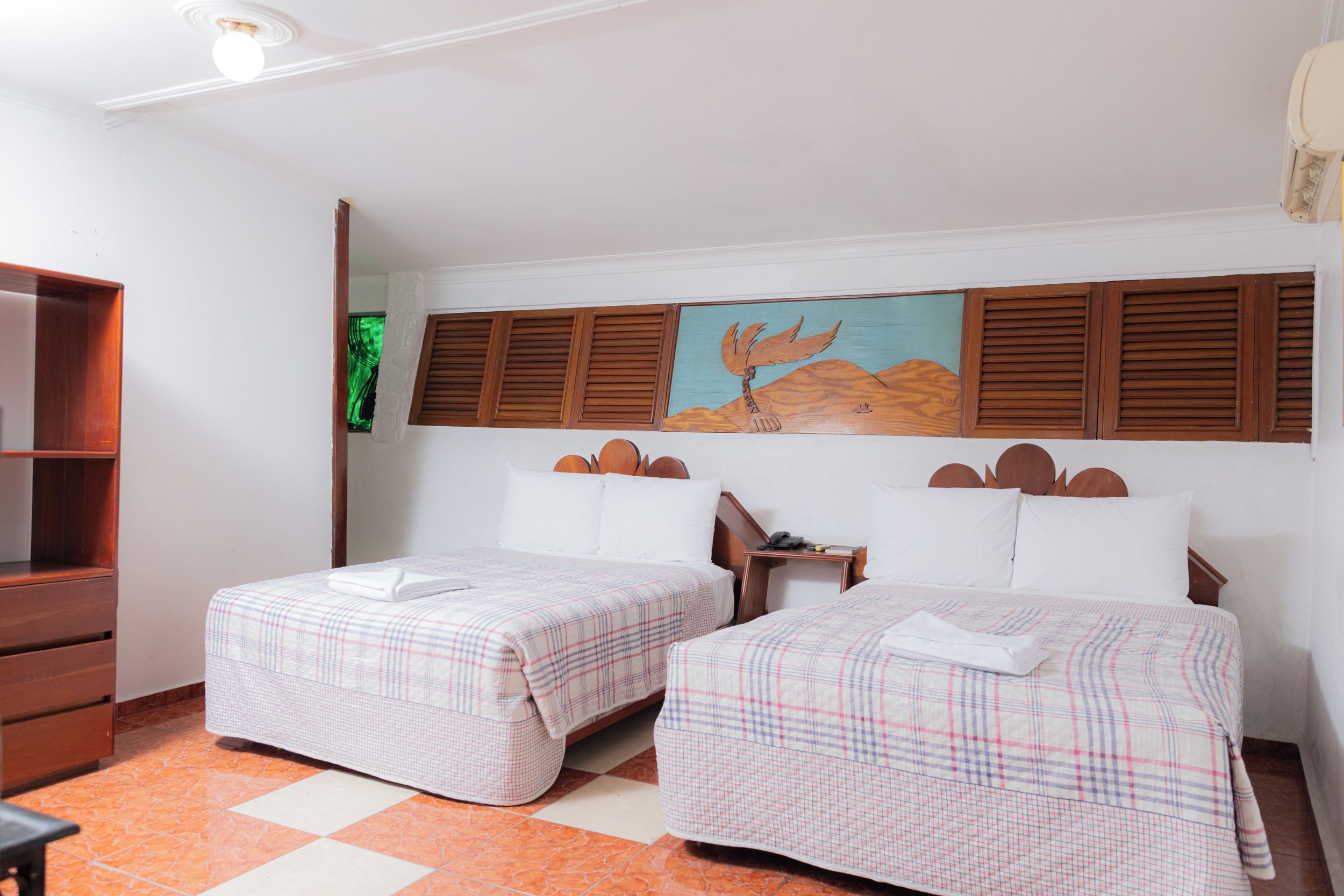 Quarto de hotel com duas camas, cabeceira de madeira, um quadro, escrivaninha e guarda-roupa.