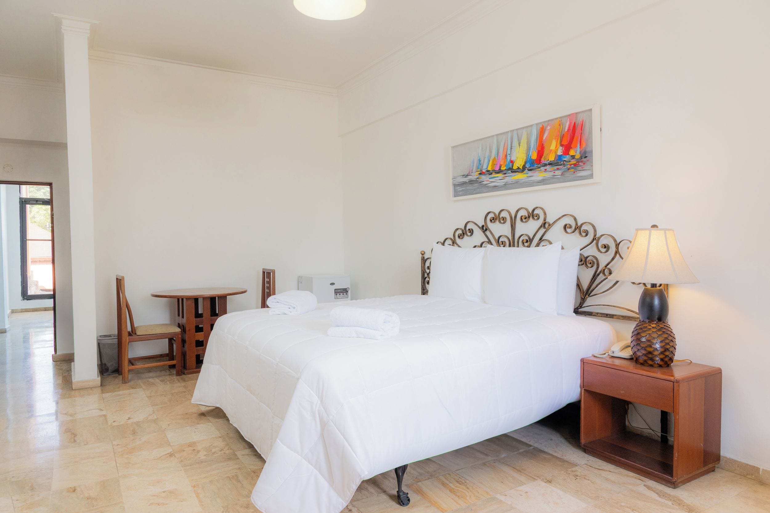 A hotel room with a bed, a bedside table with a lamp, a chair, a small table, and a painting on the wall.