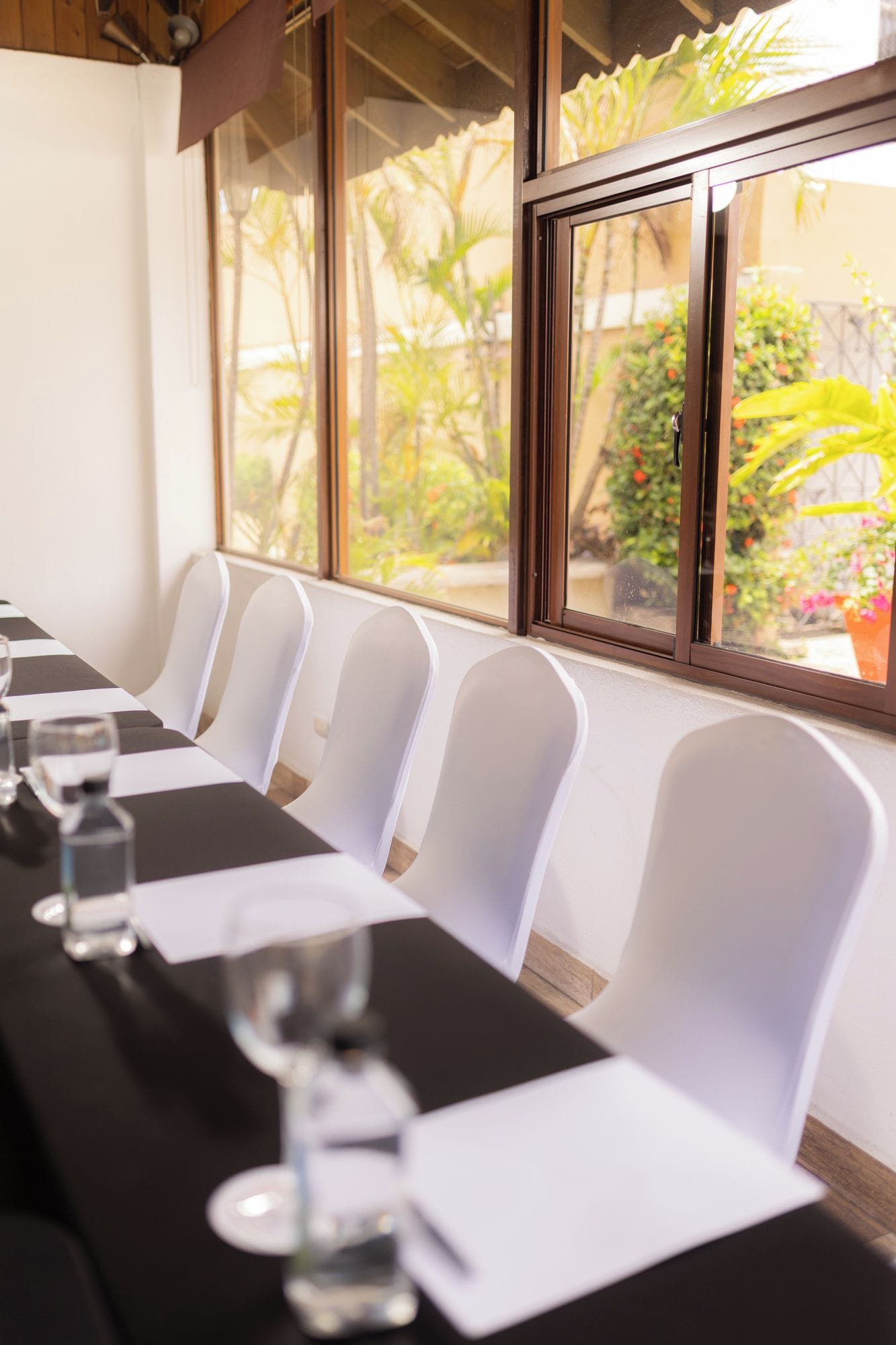 A conference room with a long table, white chairs, and a view of greenery through large windows.
