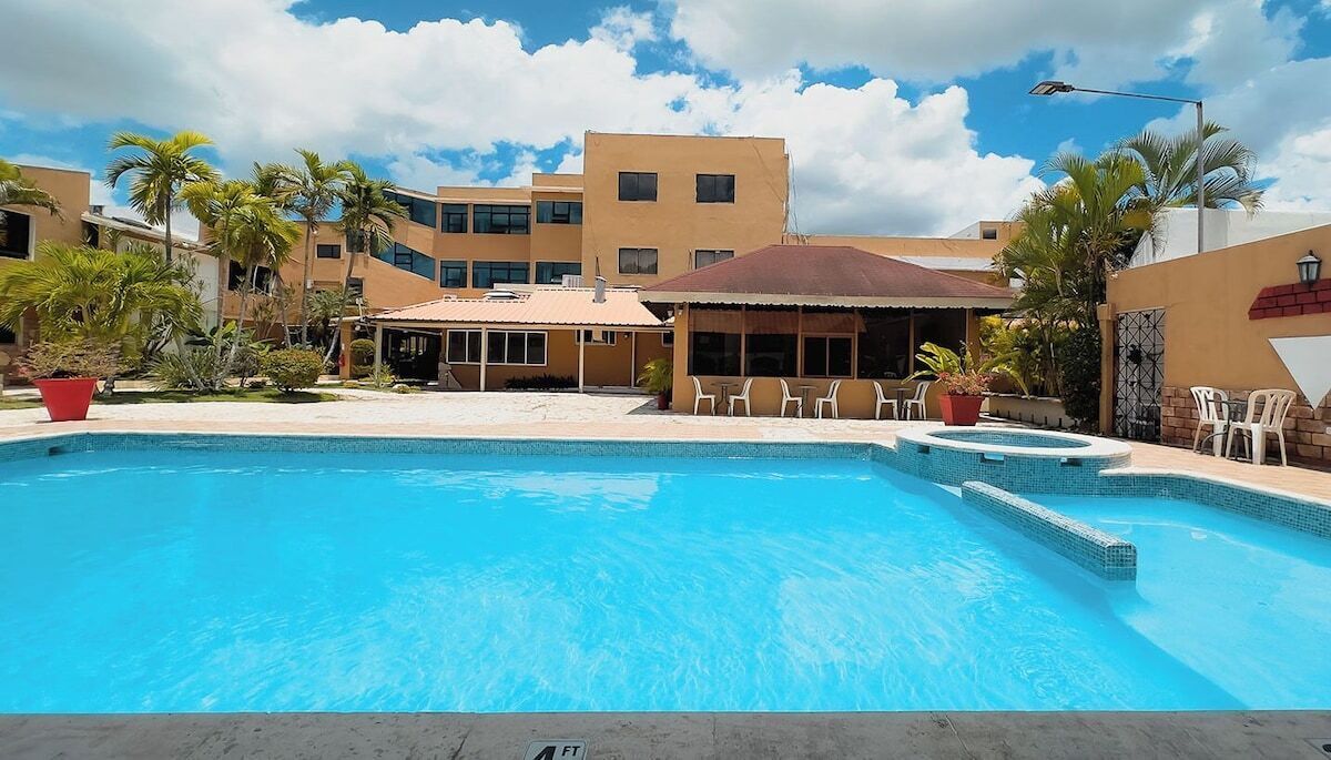 Una piscina con borde de azulejos, rodeada de palmeras y un edificio de techo plano.