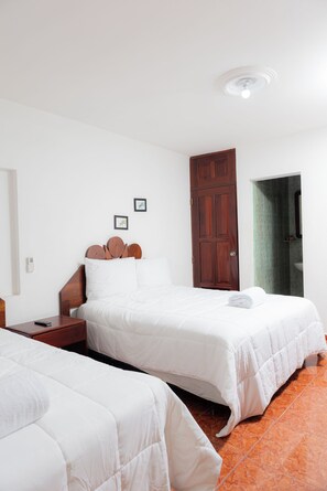 Habitación de hotel con dos camas, cabecero de madera, mesita de noche y un reloj despertador en la pared, además de una puerta que da a un baño.