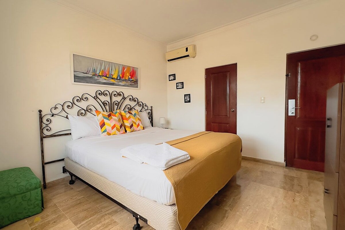 A bedroom with a bed, a green bench, a painting, and a door.