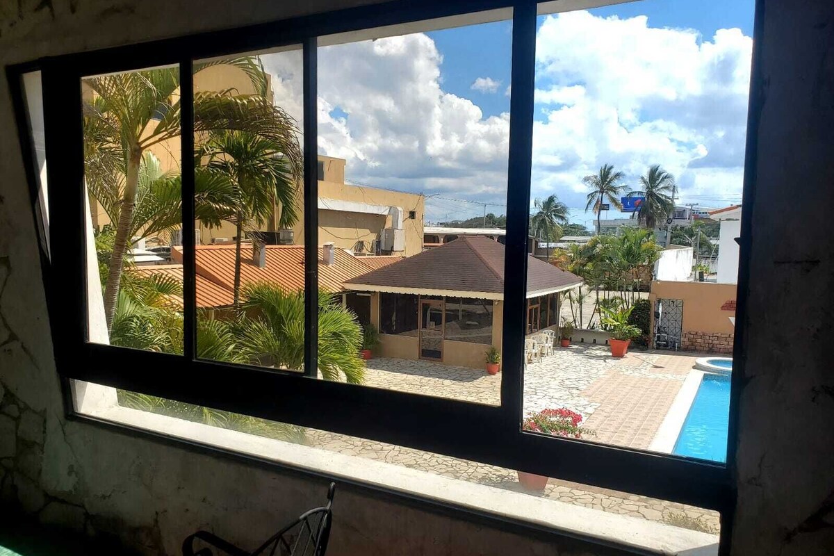 Una ventana con vista a un patio que tiene piscina, palmeras y edificios.