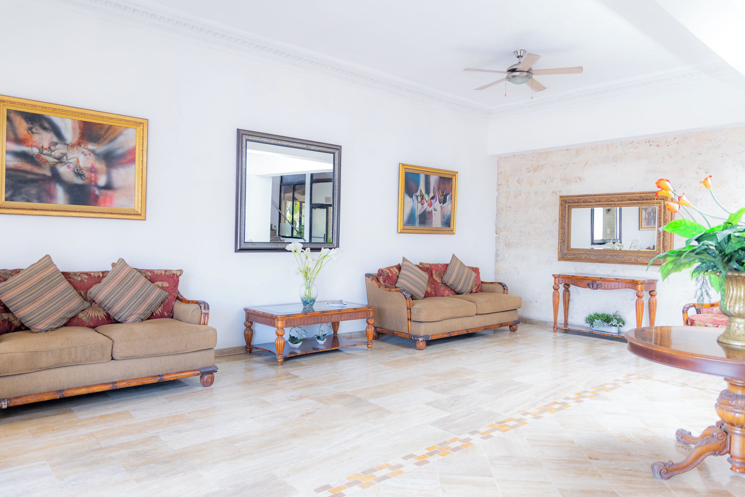 A spacious living room with a large sofa, a coffee table, and a side table. There are two framed artworks on the walls and a ceiling fan.
