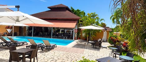 A pool area with lounge chairs, a covered seating area, and a building with a red roof.