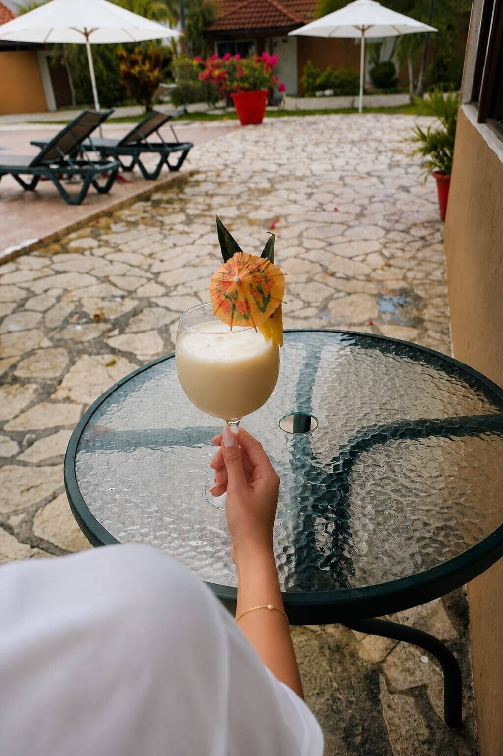 A hand holding a cocktail with a decorative umbrella on a glass table outdoors.