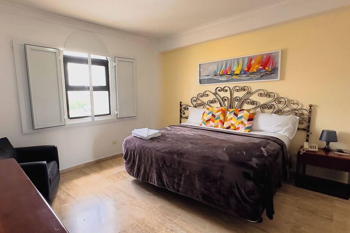 Quarto com cama, escrivaninha, cadeira, janela e um quadro na parede.