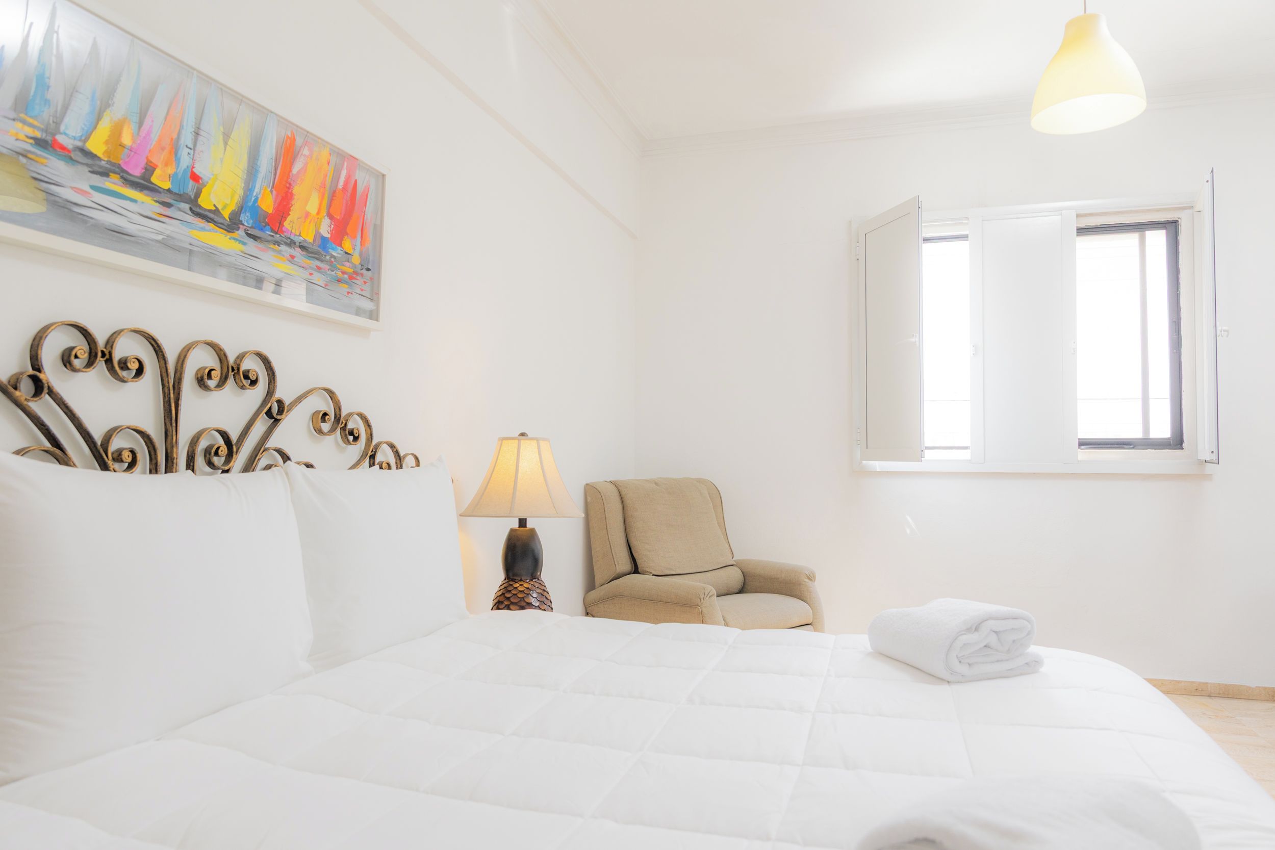 A hotel room with a bed, a chair, a lamp, a painting, and a window.
