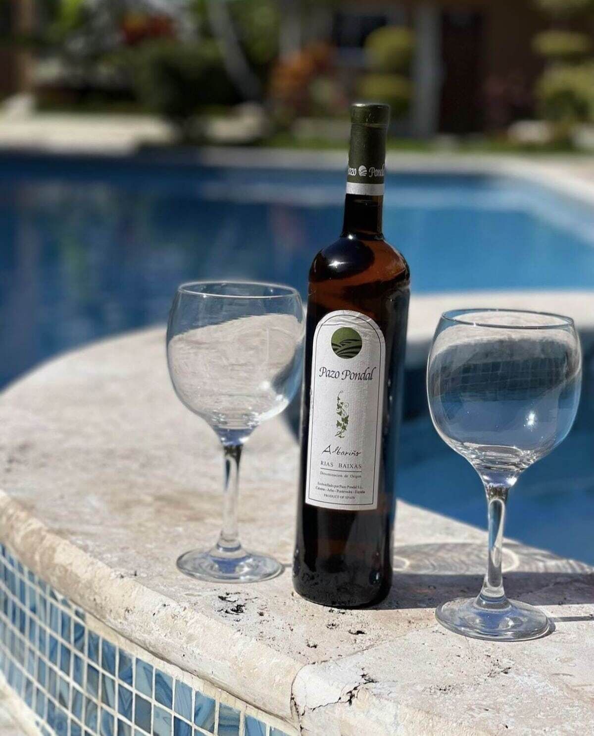 Una botella de vino Pazo Pondal y dos copas sobre una mesa junto a la piscina.