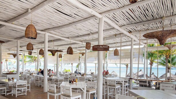 A beachside restaurant with white tables and chairs, hanging wicker lamps, and a view of the ocean. - Villa Deluxe SlowLiving Tulum CountryClub Golf PGA (Gran Bahía Príncipe)