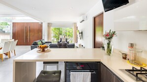 A modern kitchen with a central island, a dining area with white chairs, and a living area with a sofa and a TV. - Villa Deluxe SlowLiving Tulum CountryClub Golf PGA (Gran Bahía Príncipe)