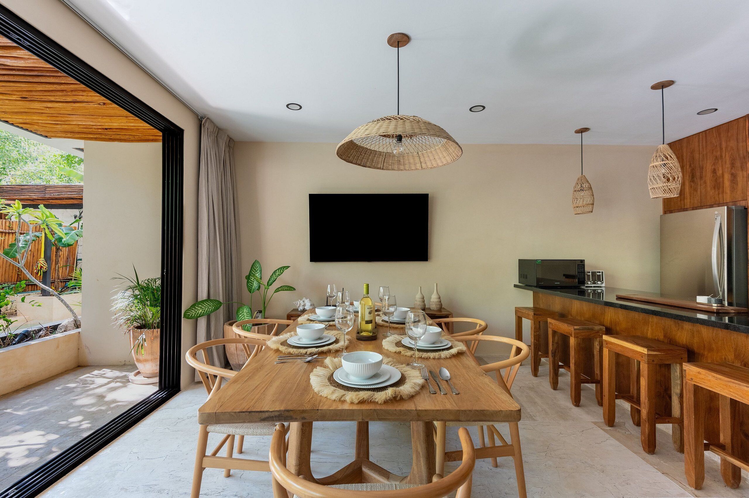 A dining area with a wooden table set for six, surrounded by wooden chairs. There is a kitchen area with a refrigerator and a microwave.