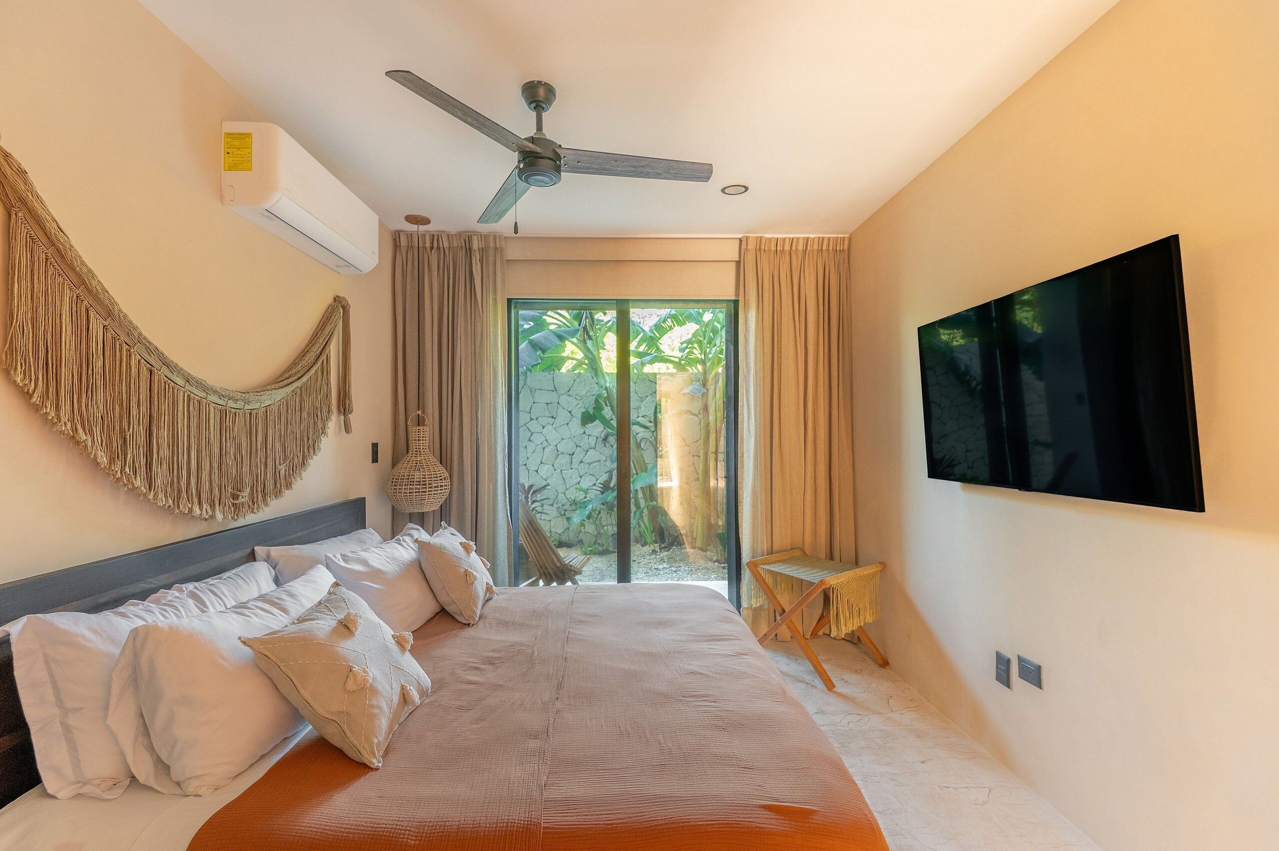 A bedroom with a bed, a ceiling fan, a TV, and a view of greenery through the glass door.