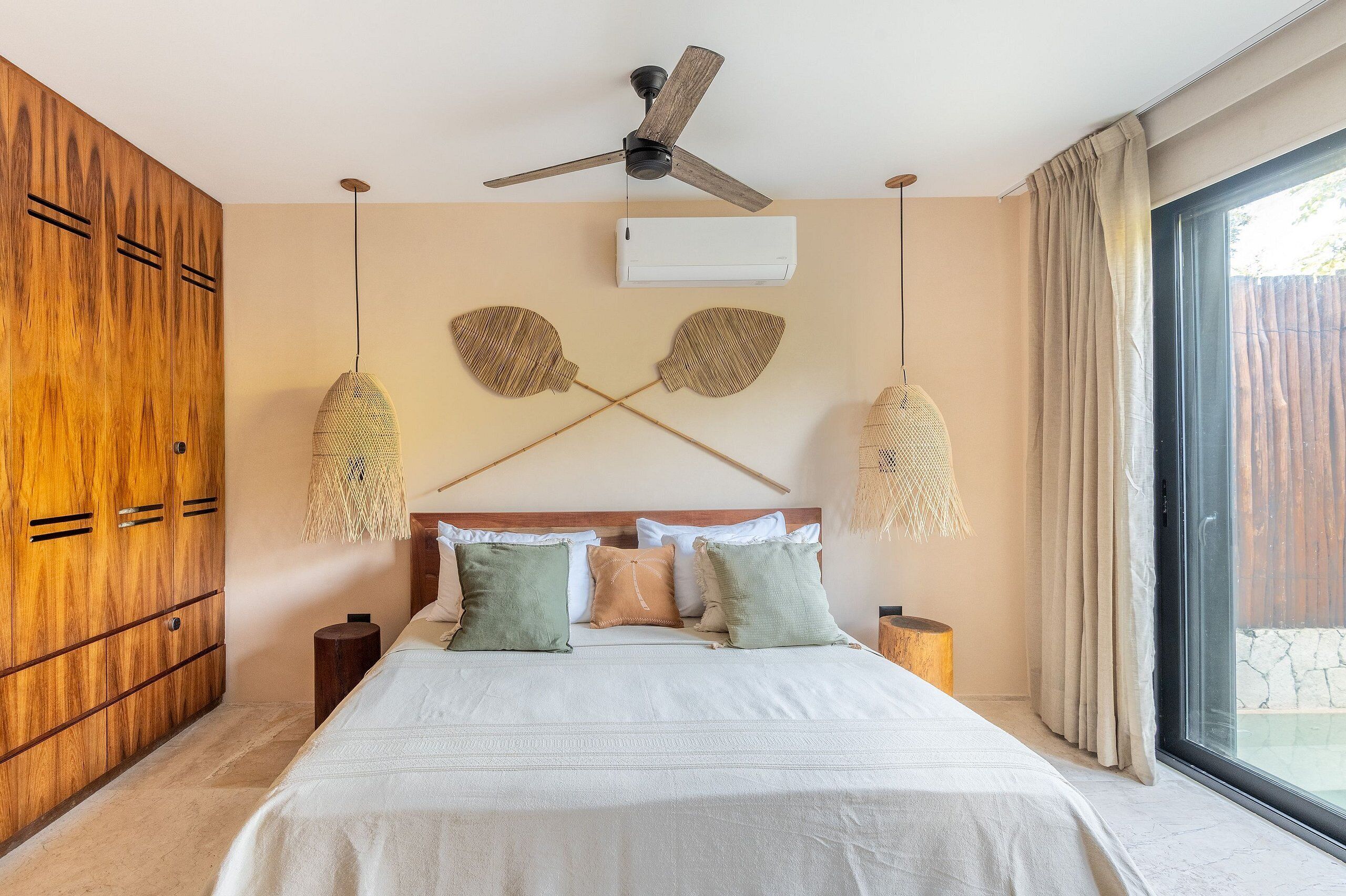 A bedroom with a bed, wooden wardrobe, ceiling fan, air conditioning unit, and a large window with curtains.