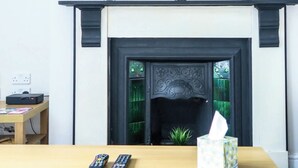 A living room with a flat-screen TV mounted above a fireplace. There are two remotes and a tissue box on a wooden table. - 5 bedroom house, free parking, near football, sleeps 8 (Liverpool)