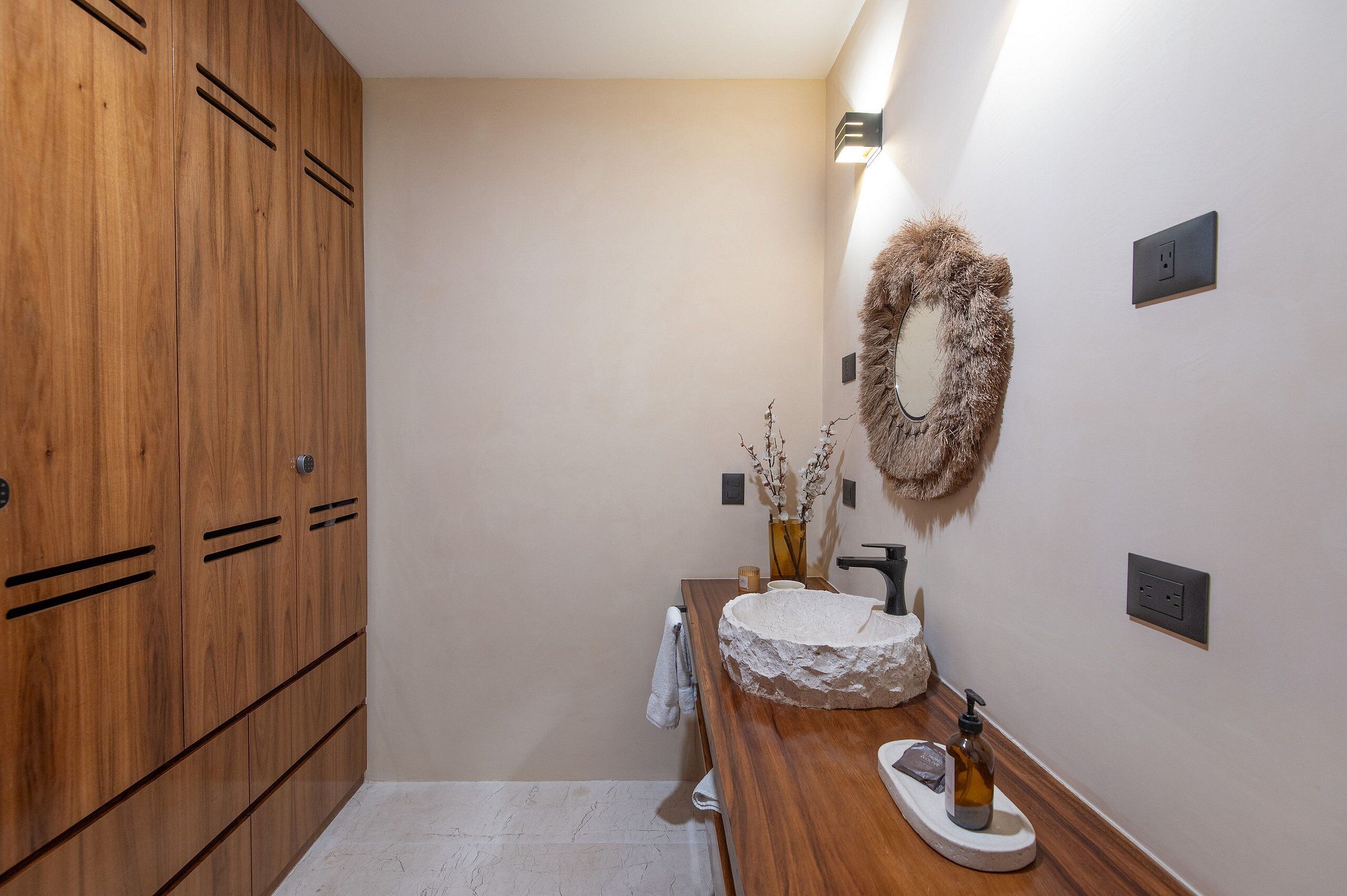 A bathroom with wooden cabinets, a white sink with a stone basin, a fluffy wall decoration, and a small vase with dried branches.