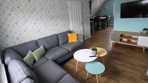 Ett modernt vardagsrum med en grå hörnsoffa, två små runda bord, en platt-TV och en vägg med ett mönster.