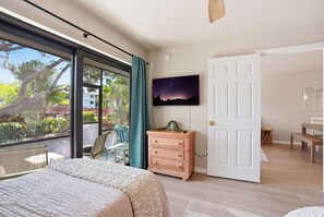 A bedroom with a bed, a dresser, a TV, and a balcony with a view of trees and a building. - B-114 -Totally Remodeled With Walk Out Lanai (Naples)