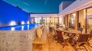 Area piscina esterna moderna con pavimento decorato, tavolo con sedie e vista sul cielo.