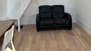 A room with a wooden floor, a black leather sofa, a white chair, and a white staircase with exposed beams. - Luxembourg Montparnasse (Paris)