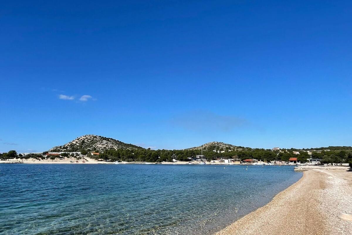 A beach with clear blue water, a sandy shore, and a hill in the background.