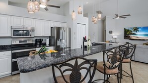 A modern kitchen with white cabinets, stainless steel appliances, and a granite countertop. There are two bar stools with woven seats and a large island with a built-in sink. - Lely Resort Single-Family Home Monthly Min Stay (Naples)