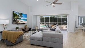 A spacious living room with a large window, a ceiling fan, and a view of a pool. - Lely Resort Single-Family Home Monthly Min Stay (Naples)