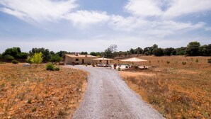 Ein Kiesweg führt zu einem Gebäude mit Außenplätzen und einem Pool, umgeben von trockenem Gras und Bäumen.