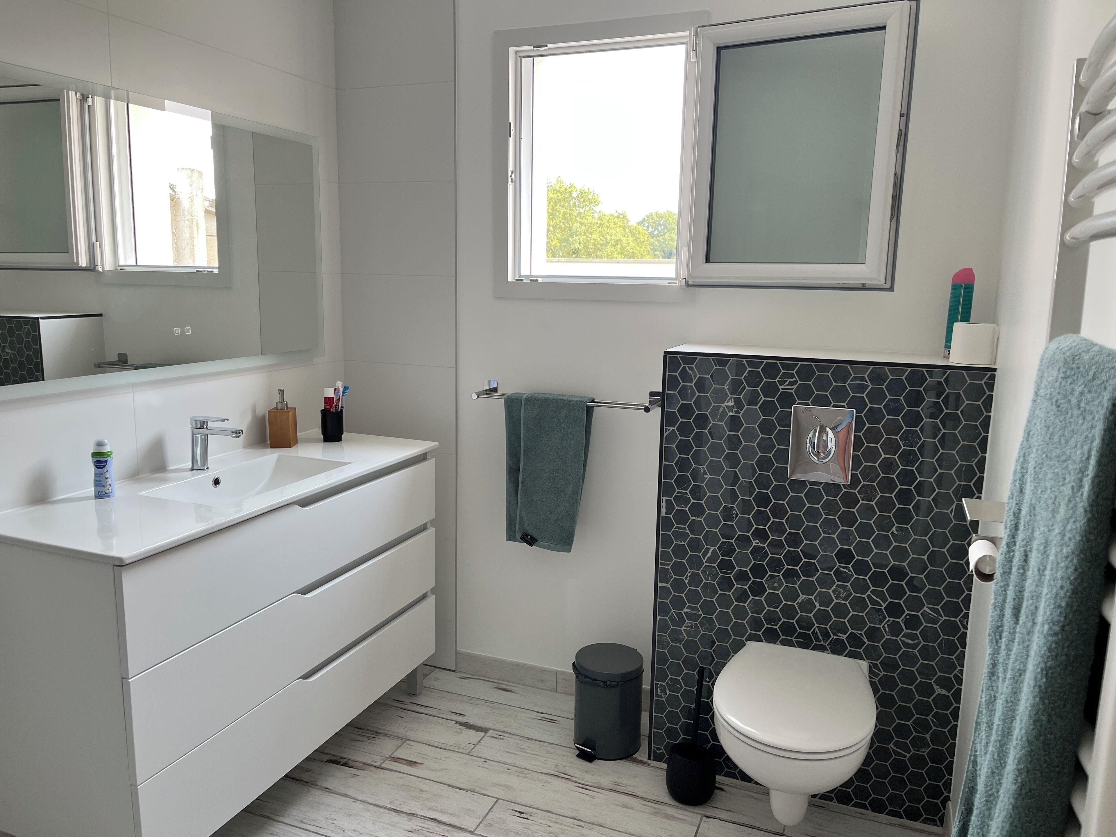 A modern bathroom with a white vanity, a black tiled shower wall, a toilet, and a window providing natural light.