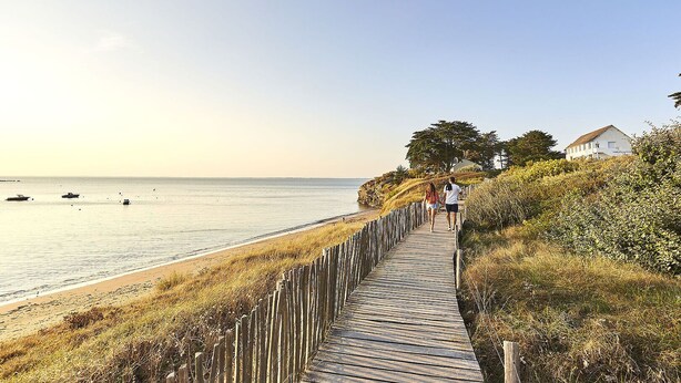 Un paseo marítimo de madera junto a la playa, con vista despejada al mar y una casa al fondo.