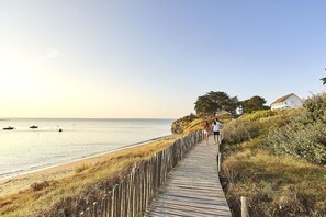 Ein hölzerner Steg entlang eines Strandes mit freiem Blick auf das Meer und einem Haus im Hintergrund.