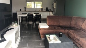 Una sala de estar moderna con un sofá marrón, una mesa de centro y un televisor. Se observa una cocina al fondo con una mesa de comedor y sillas.