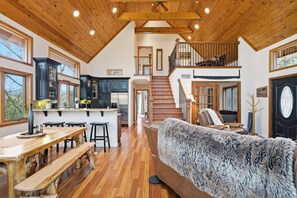 A spacious interior with a wooden ceiling, a dining area with a wooden table and benches, a cozy living room with a fur-covered sofa, and a kitchen with dark cabinets. - Pet-Friendly Home Near Vogel State Park (Blairsville)