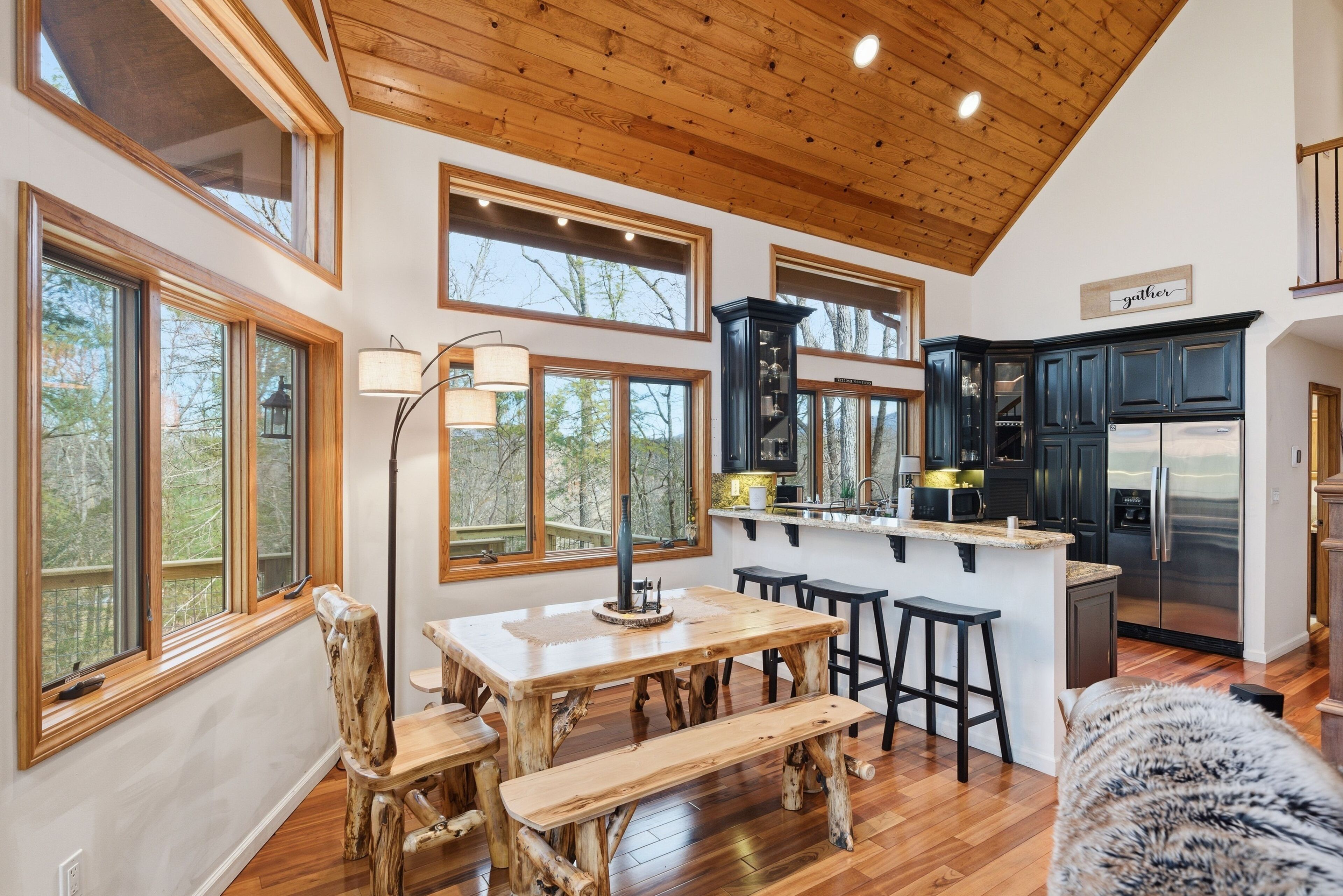 A spacious kitchen with wooden cabinetry, a central island with stools, and large windows offering a view of the outdoors.