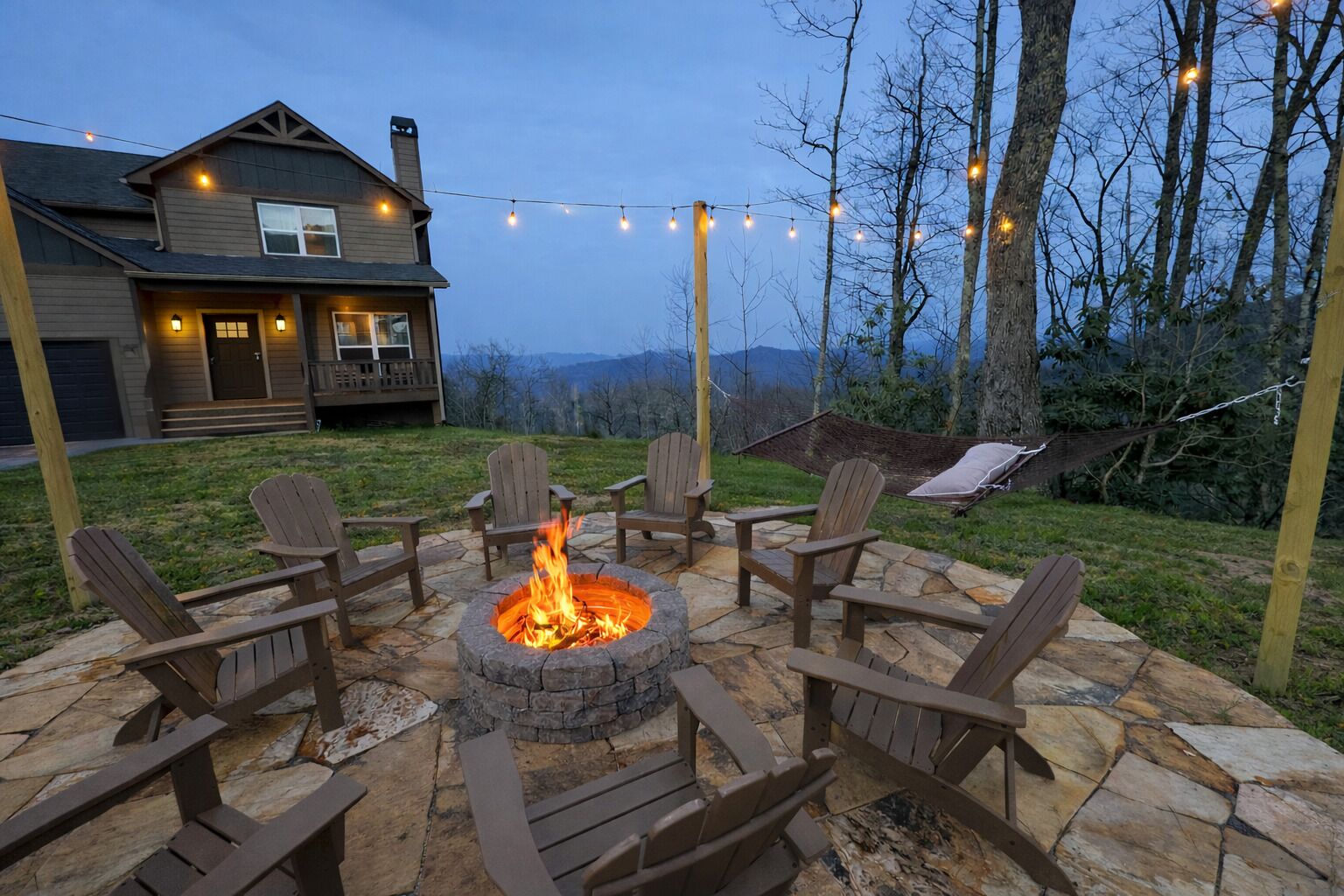 A stone fire pit with chairs, a hammock, and string lights in a backyard with a house and mountains in the background.