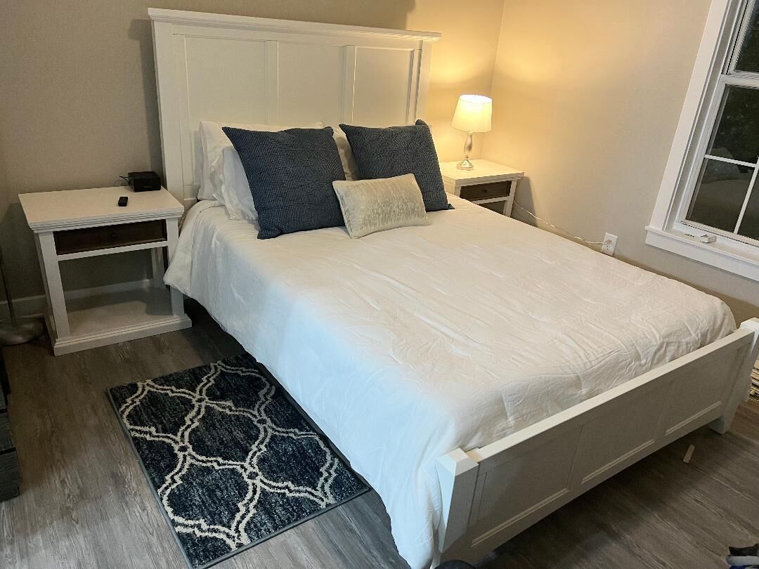 A neatly made bed with a white headboard and pillows, a bedside table with a lamp, and a patterned rug on a wooden floor.