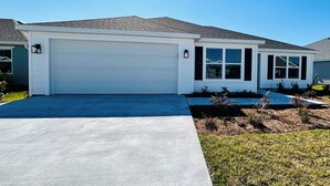A single-story house with a white garage door, a covered entryway, and a small front yard with landscaping. -  Sun-Filled Brand-New Edenfield Designer Home (Sumterville)
