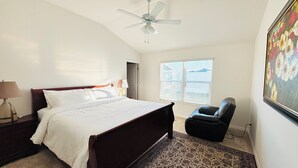 A bedroom with a bed, a ceiling fan, a chair, a window with blinds, and a painting on the wall. -  Sun-Filled Brand-New Edenfield Designer Home (Sumterville)