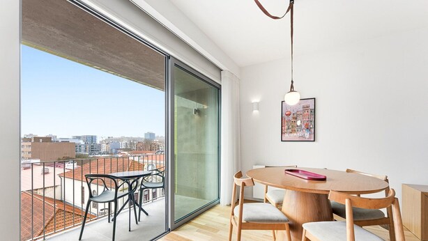 A modern dining area with a round wooden table and chairs, a glass partition, and a city view. - GuestReady - Seaside retreat - 4D (Matosinhos)